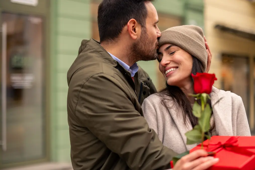 Un couple s'offrent des cadeaux à l'occasion de la Saint-Valentin. L'homme porte une veste verte et serre dans ses bras sa partenaire, qui porte un manteau beige. Ils tiennent une rose rouge et font leur cadeau en plein air, dans un environnement urbain.